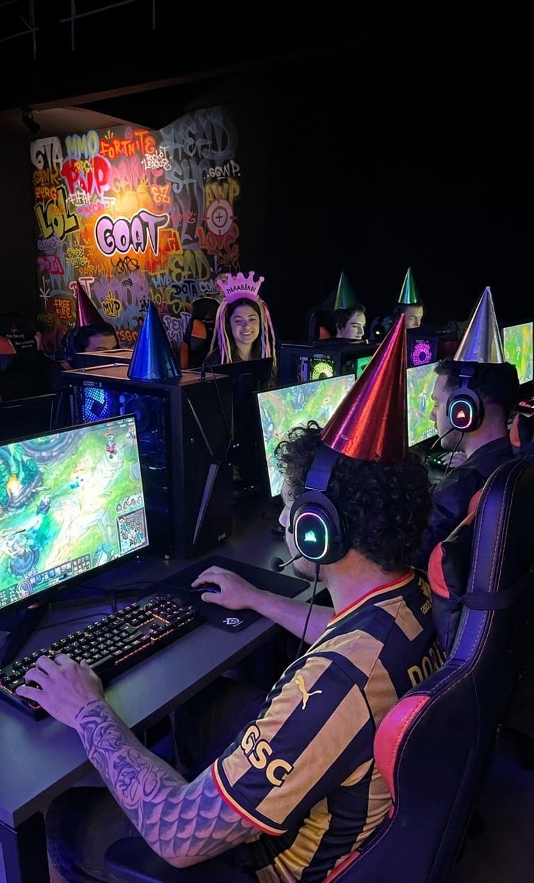 Grupo comemorando aniversário gamer na GOAT com chapéus de festa, jogando nos PCs em frente à parede grafitada
