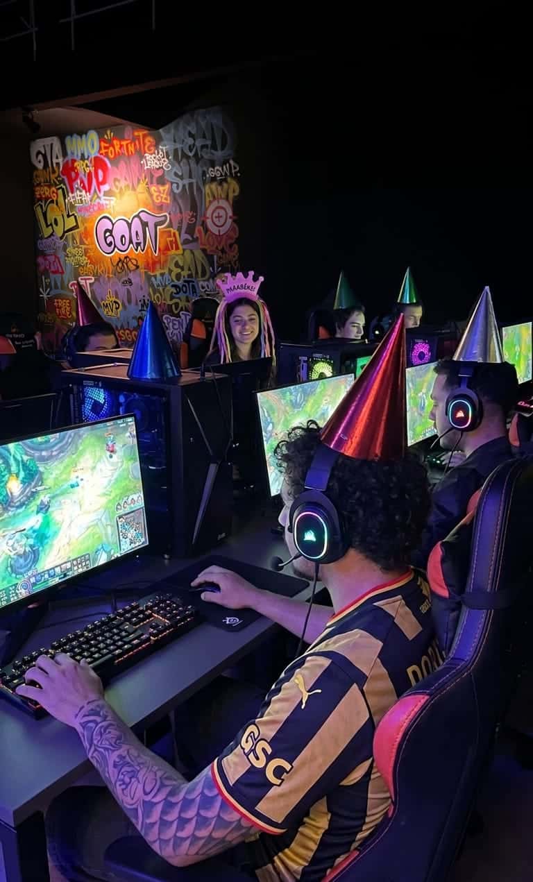 Grupo comemorando aniversário gamer na GOAT com chapéus de festa, jogando nos PCs em frente à parede grafitada