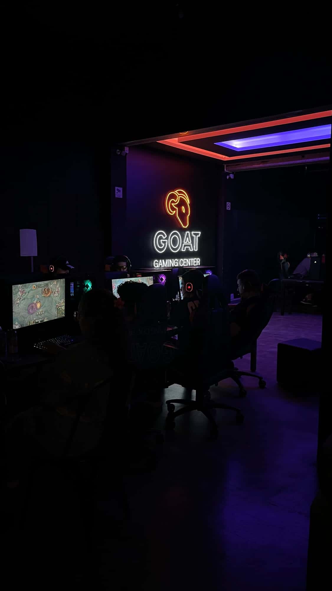 Salão da GOAT Savassi à meia-luz com PCs gamer e o logo GOAT na parede